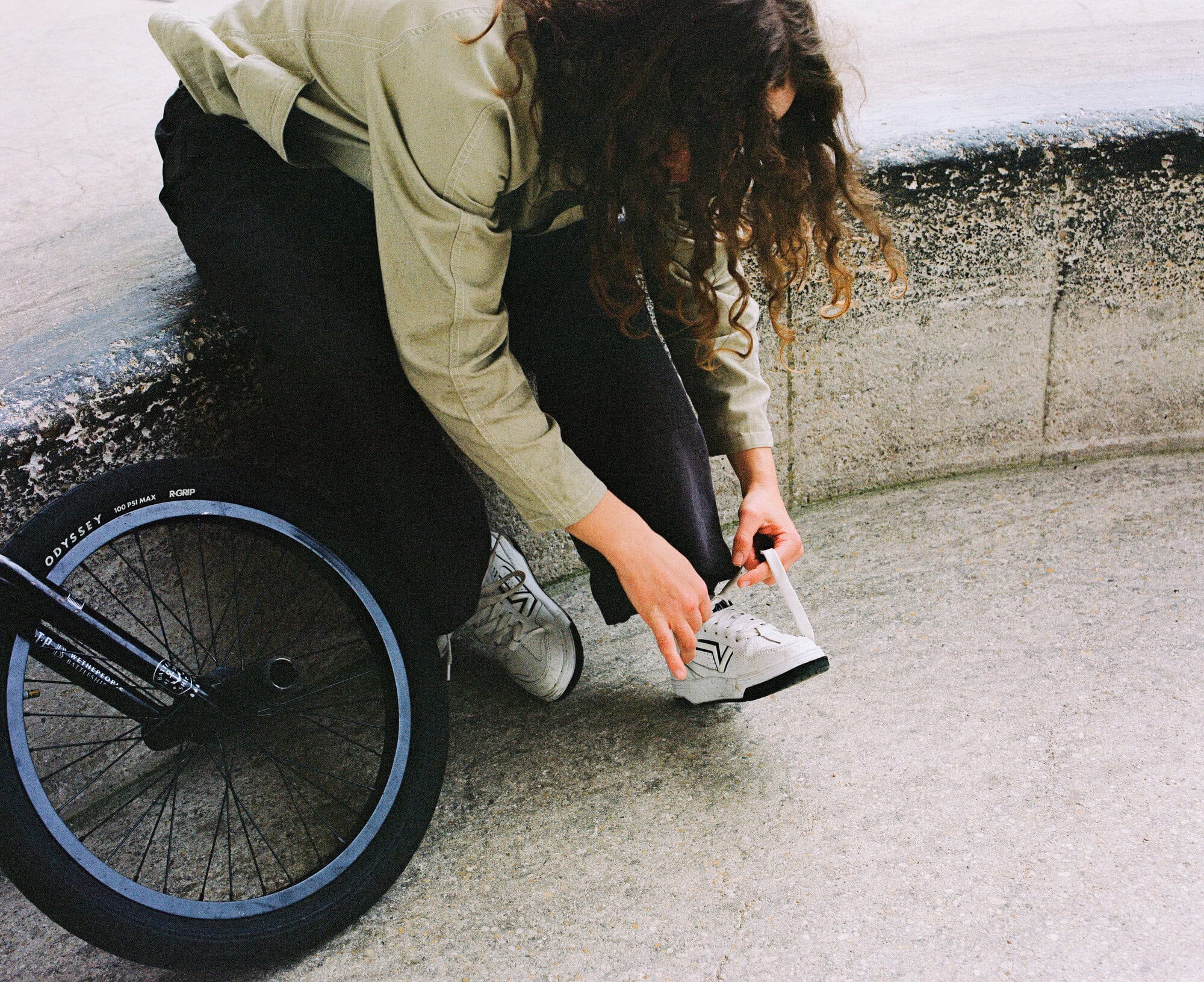 Young woman with curly hair kneeling next to a BMX bike on concrete, tying her shoelace. The image is shot in a medium shot.