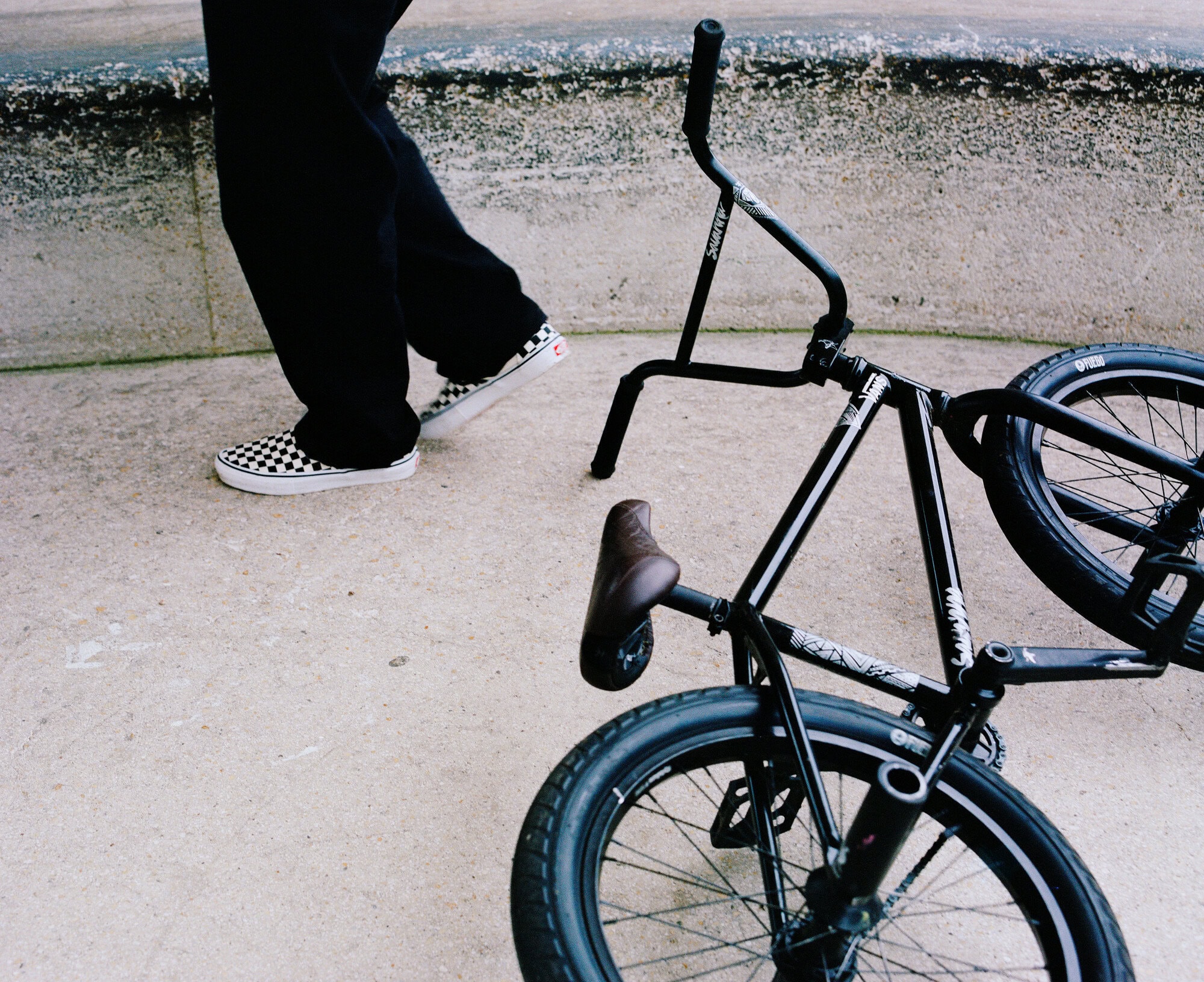 BMX bicycle on the ground next to a person walking wearing black pants and checkered Vans sneakers on pavement outside.