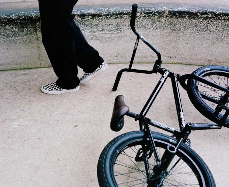 BMX bicycle on the ground next to a person walking wearing black pants and checkered Vans sneakers on pavement outside.