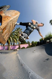 Skater performing a trick above a concrete bowl with palm trees and a large metal excavator bucket in the background under clear blue sky