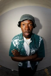 Black man wearing a green patterned button-down shirt, white t-shirt, bucket hat, and gold chain against a light grey wall