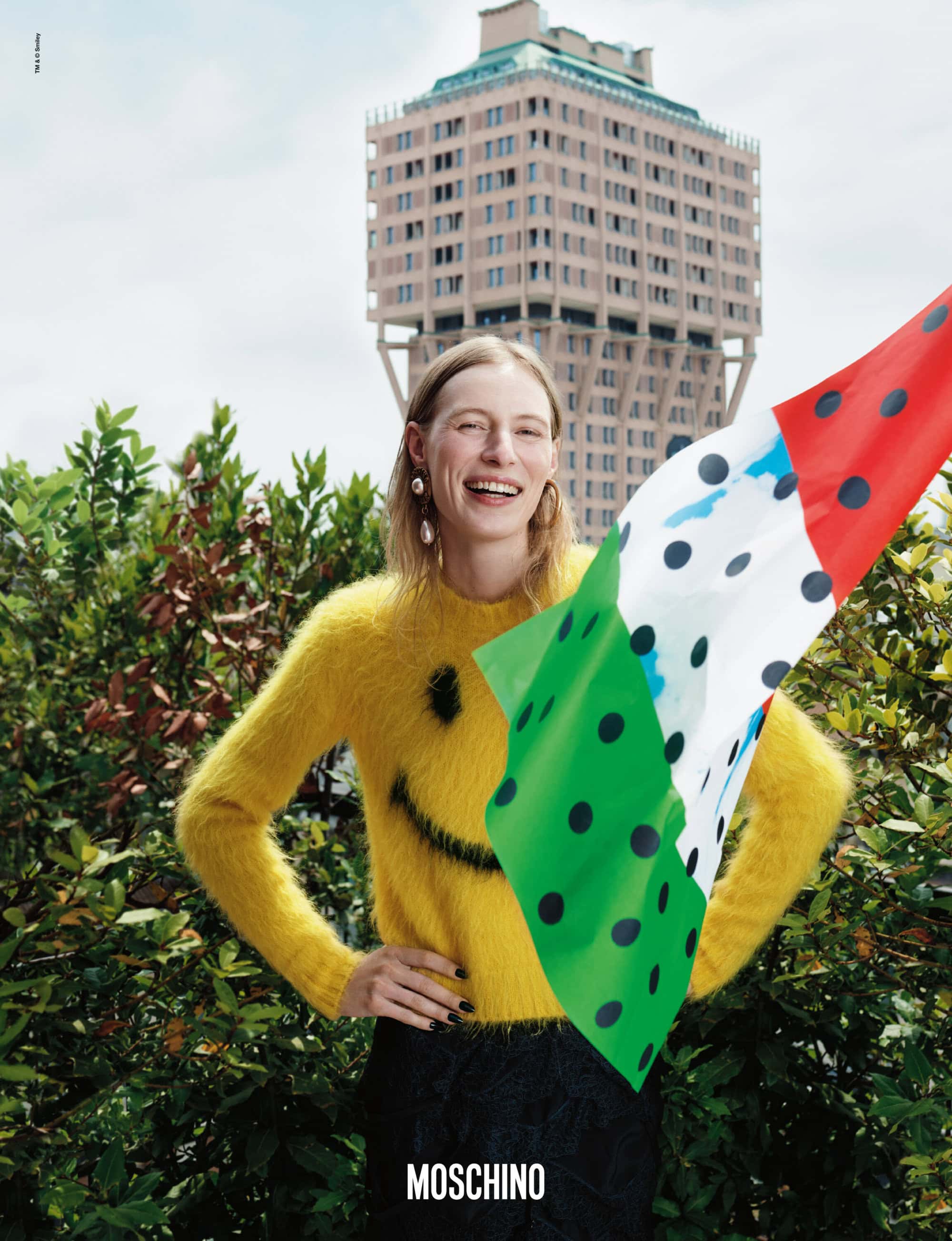 Fashion editorial shot of a smiling woman in a yellow smiley-face sweater, holding a polka-dot fabric, with a building in the background.
