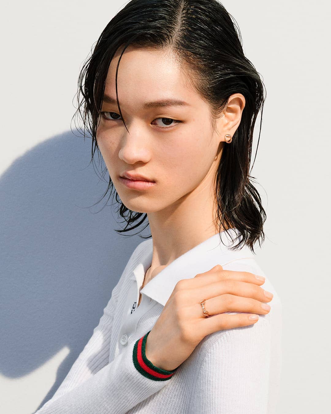 Young Asian woman in a white collared shirt with red and green sleeve details, a gold ring, and slicked-back dark hair poses indoors.
