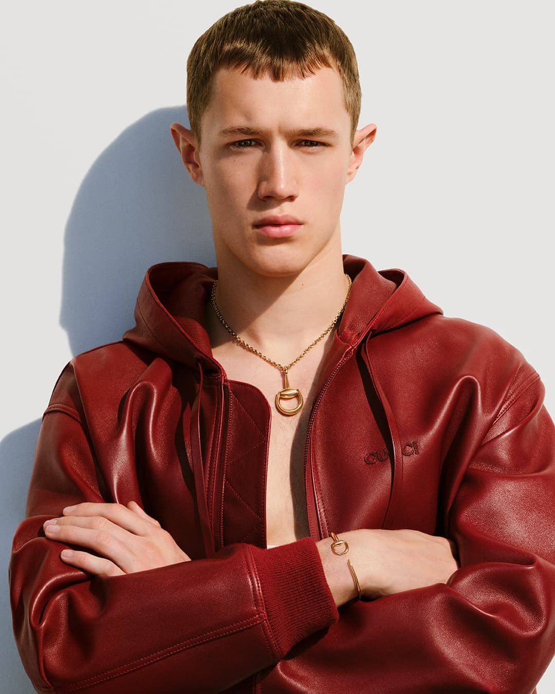 Portrait of a fair-skinned young adult male wearing a red leather hooded jacket, gold chain necklace, and bracelet against a white background.