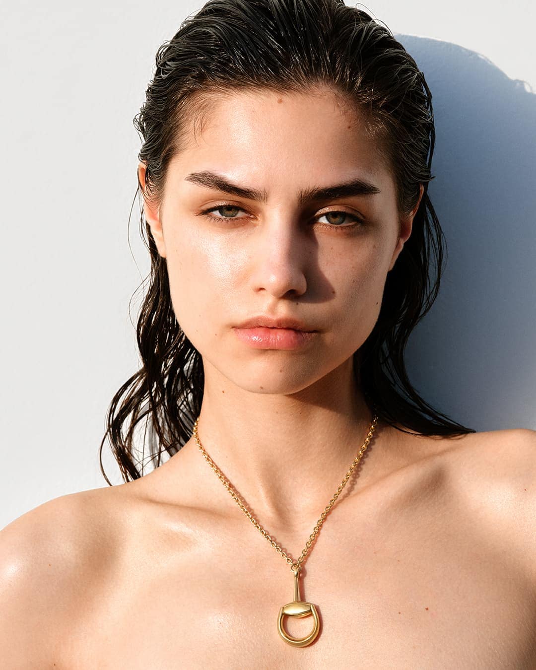 Close-up of a young woman with wet dark hair wearing a gold pendant necklace against a plain white background in studio lighting.