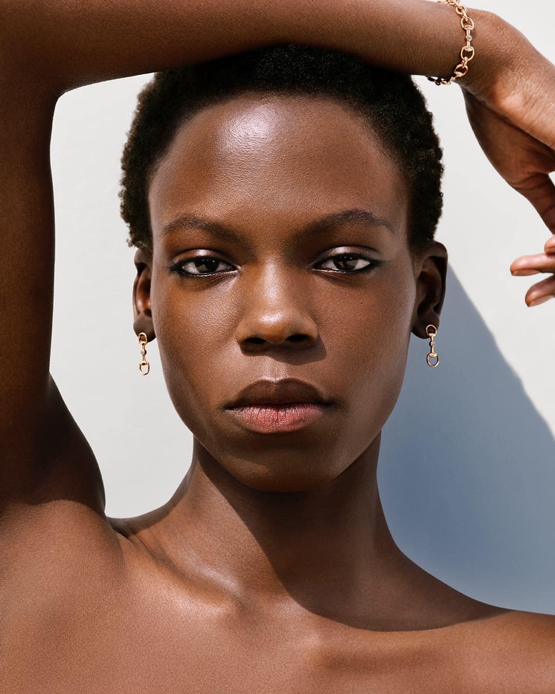 Close-up portrait of a model with short dark hair wearing gold horsebit earrings and bracelet against a plain white background