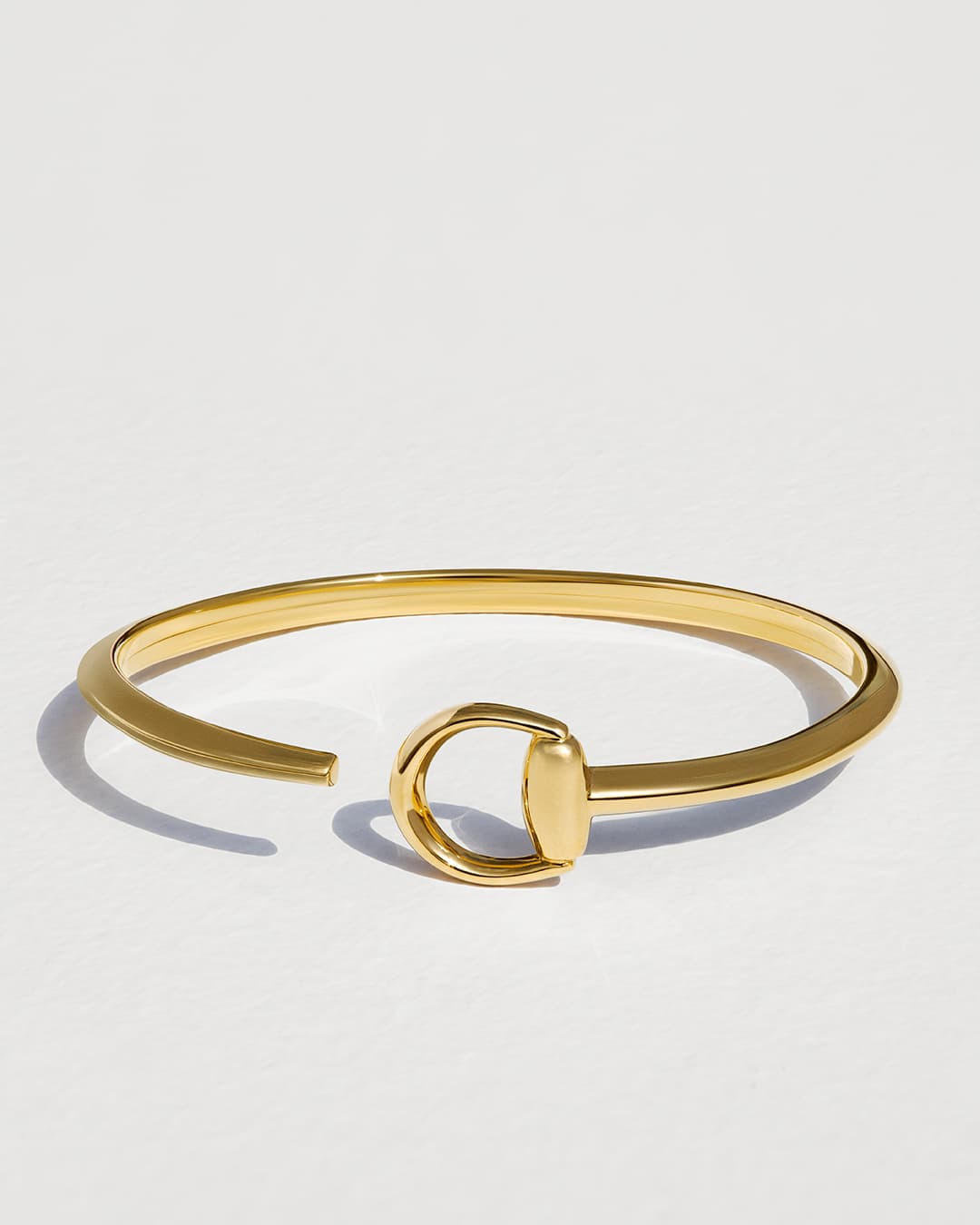 Gold-toned metal bracelet with a horseshoe-shaped closure on a white surface in a studio setting with soft lighting.