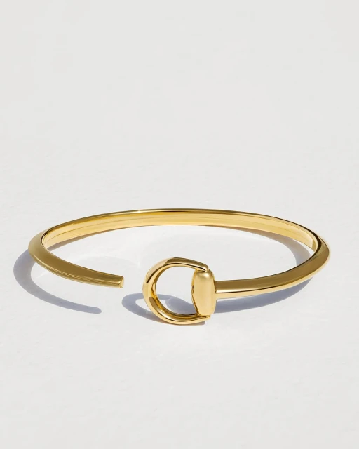 Gold-toned metal bracelet with a horseshoe-shaped closure on a white surface in a studio setting with soft lighting.