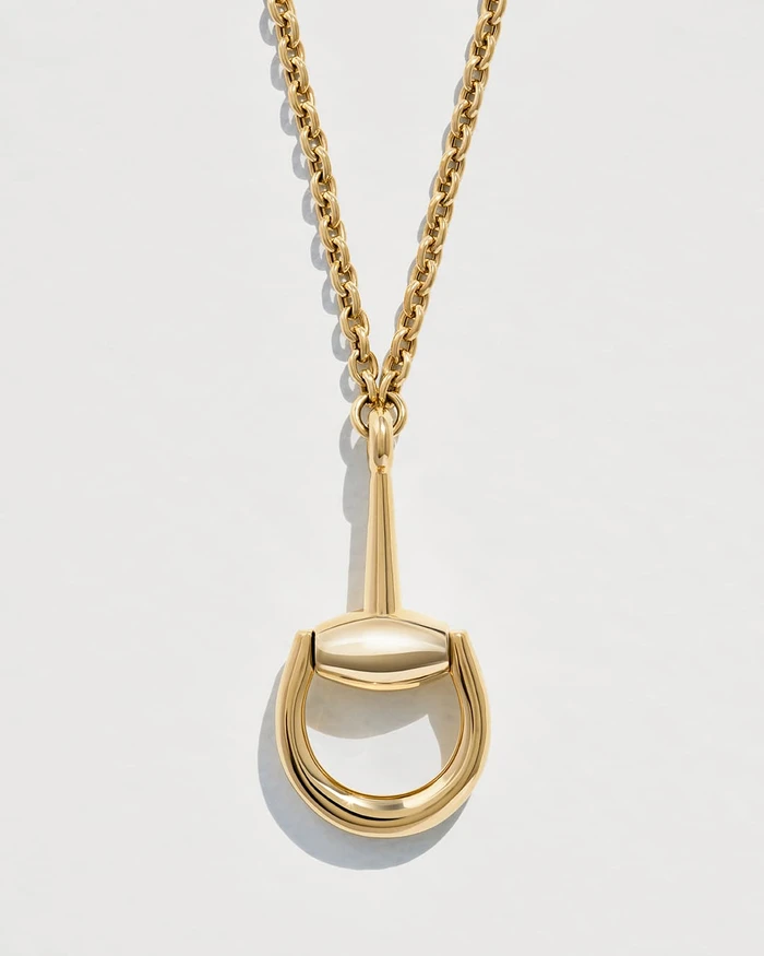 Close-up of a polished gold horsebit pendant necklace centered against a smooth white studio background.