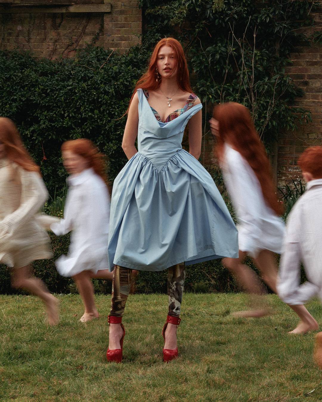 A red-haired woman poses in a blue dress with a full skirt, set against a green lawn and hedges, surrounded by blurred figures in motion.