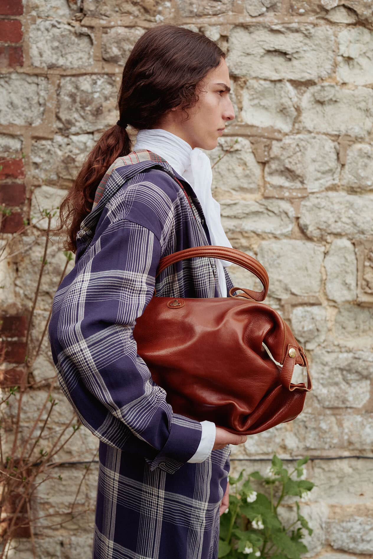 Model in plaid coat holding a brown leather bag against a stone building exterior wall