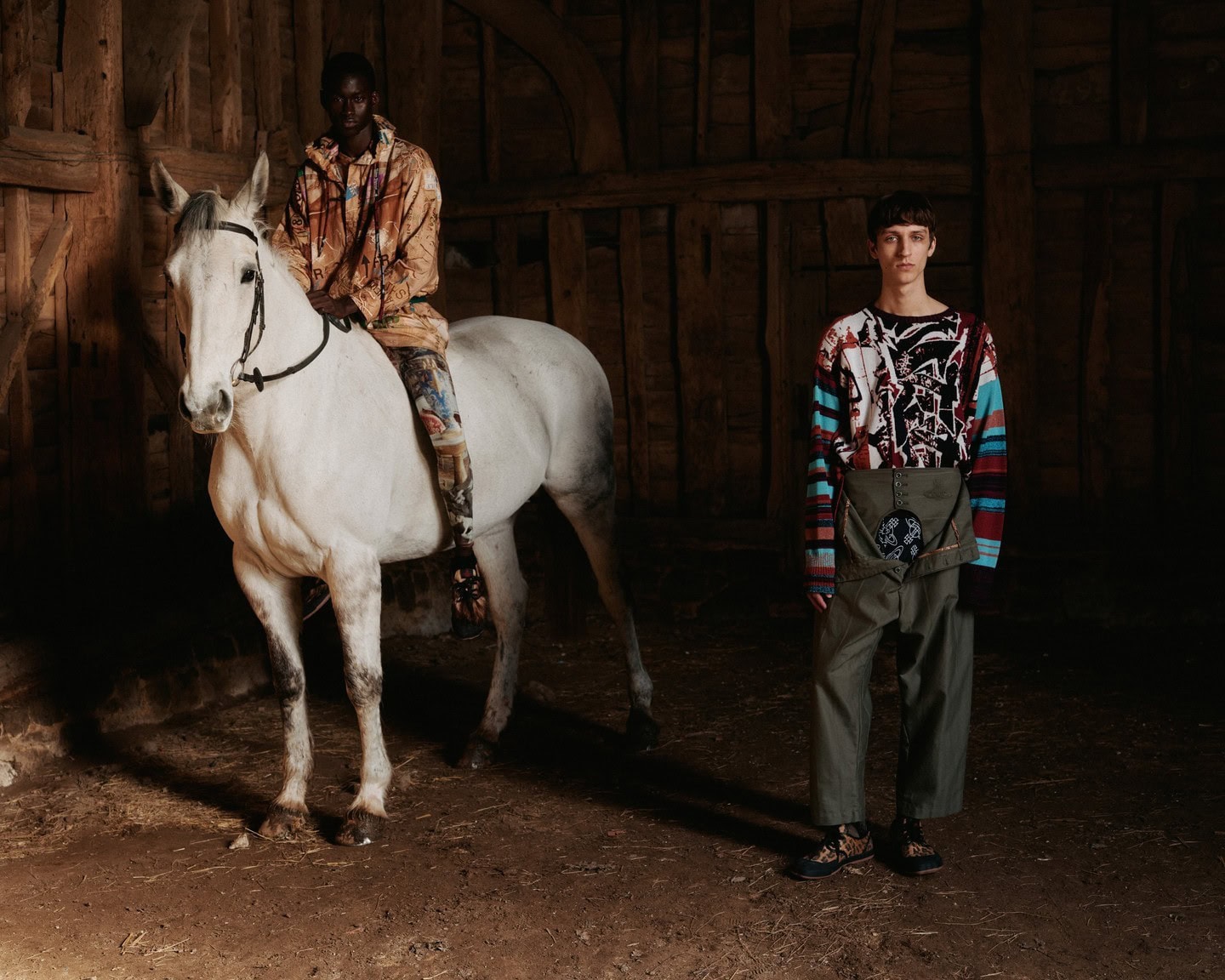 A light-skinned man stands next to a white horse being ridden by a dark-skinned male indoors, illuminated by natural light in an editorial style.