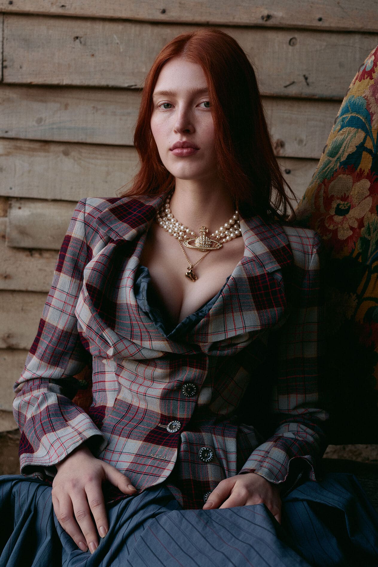 Red-haired woman in a plaid blazer with pearl necklace. She is sitting, looking at the camera, against a rustic wooden wall.