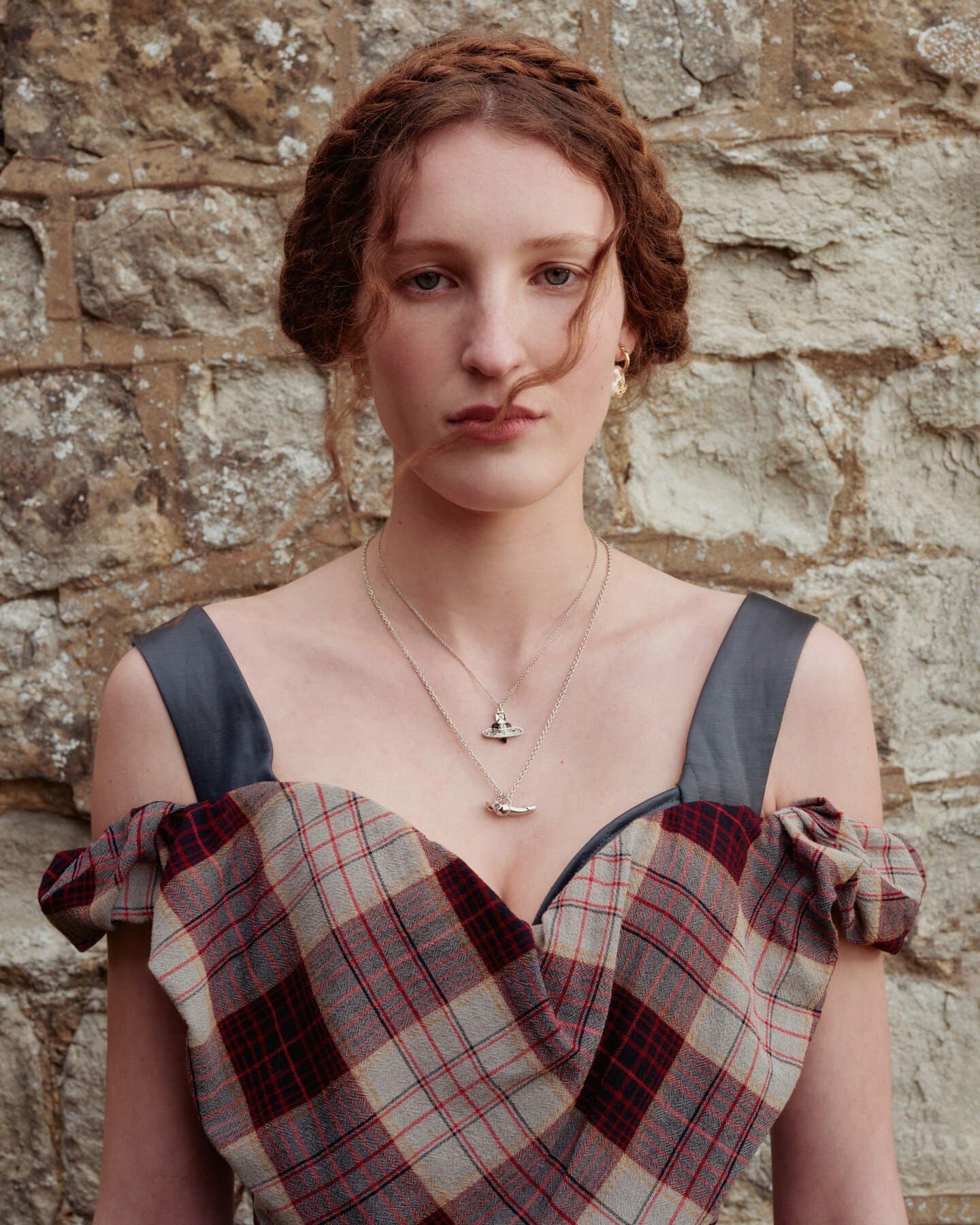 Portrait of a red-haired woman with a serious expression wearing a plaid dress and necklaces against a stone wall background.