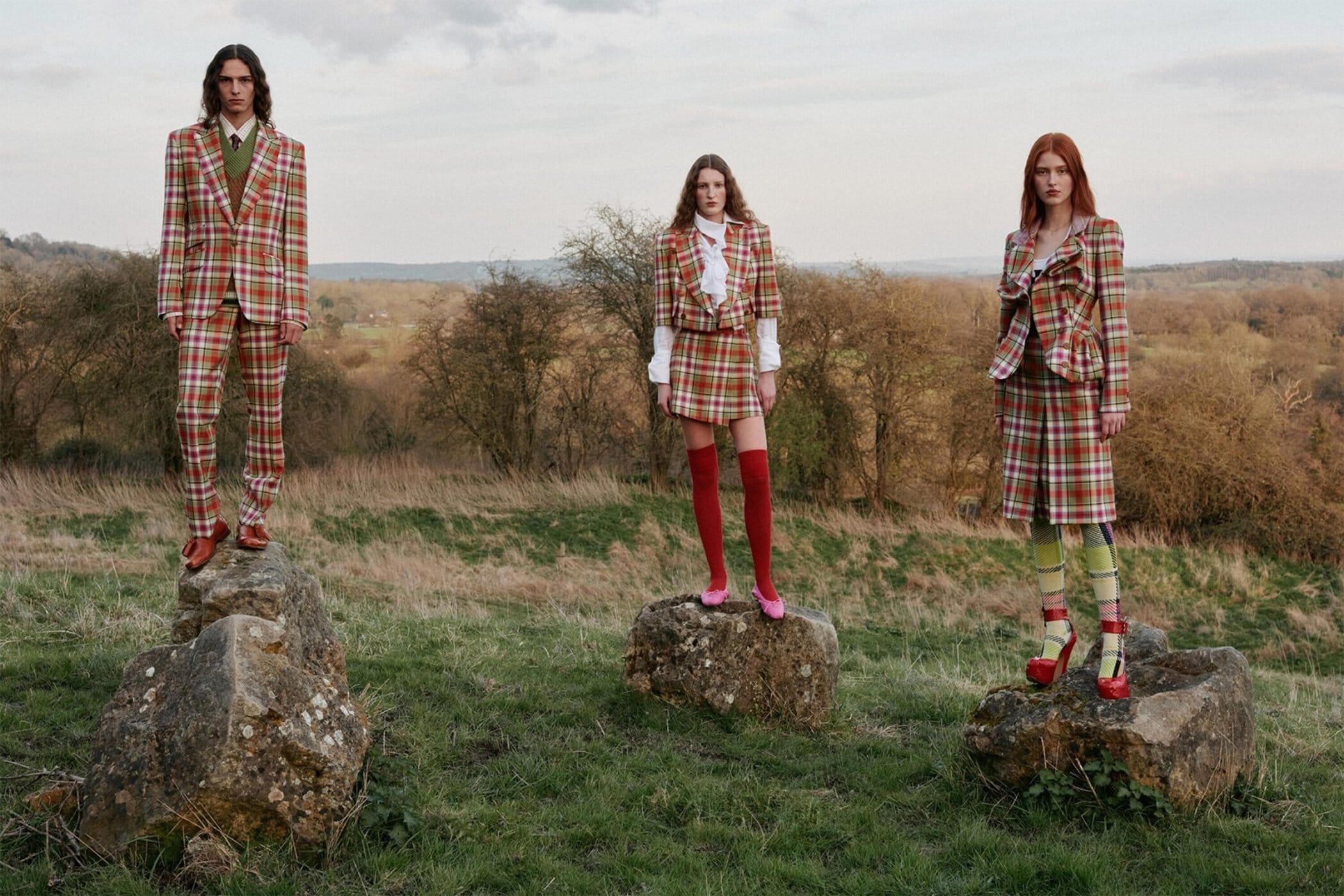 Three fashion models in plaid outfits stand on rocks in a grassy field with a backdrop of rolling hills and a light sky.