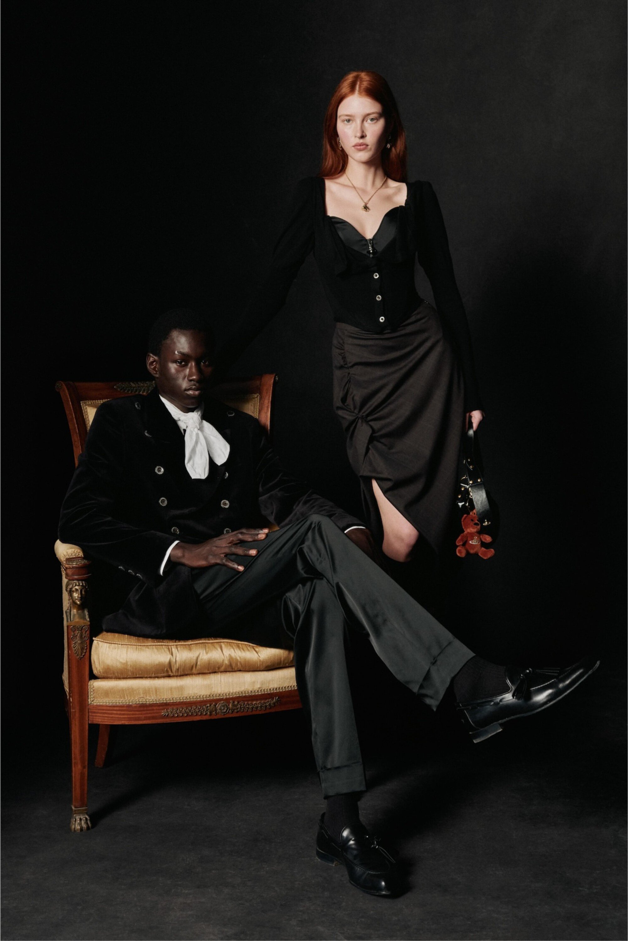 A stylish couple poses in a photo studio. The man is seated in an ornate chair, and the woman stands, wearing elegant dark attire.