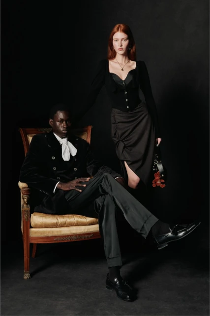 A stylish couple poses in a photo studio. The man is seated in an ornate chair, and the woman stands, wearing elegant dark attire.