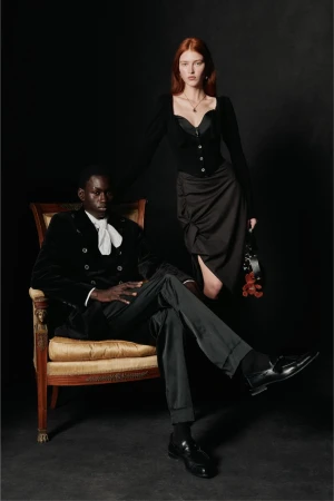 A stylish couple poses in a photo studio. The man is seated in an ornate chair, and the woman stands, wearing elegant dark attire.