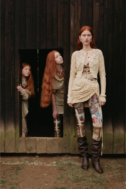 Three red-haired women pose in a rustic outdoor setting. One stands in front, wearing a knit outfit and boots, while two others stand in a wooden doorway.