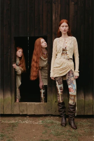 Three red-haired women pose in a rustic outdoor setting. One stands in front, wearing a knit outfit and boots, while two others stand in a wooden doorway.