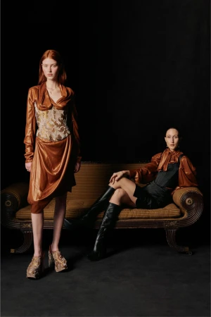 Two models wearing Vivienne Westwood clothing, one standing in a copper-toned dress and the other seated on a vintage sofa in black.