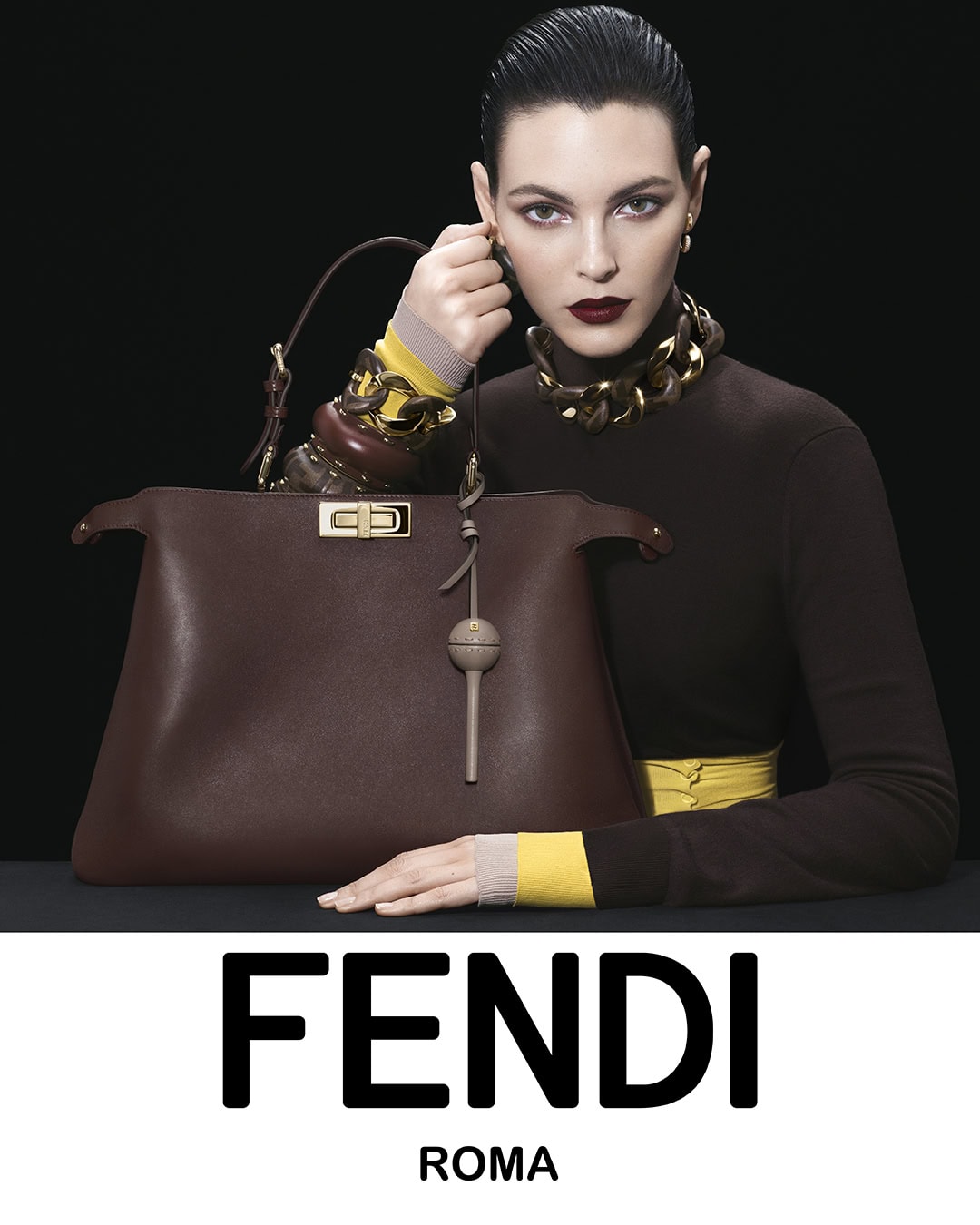 Fashion ad featuring a brunette woman wearing a brown turtleneck and a gold necklace, holding a Fendi handbag with the brand's logo.