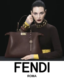 Model holding a large dark brown leather handbag wearing a brown turtleneck sweater against a black background