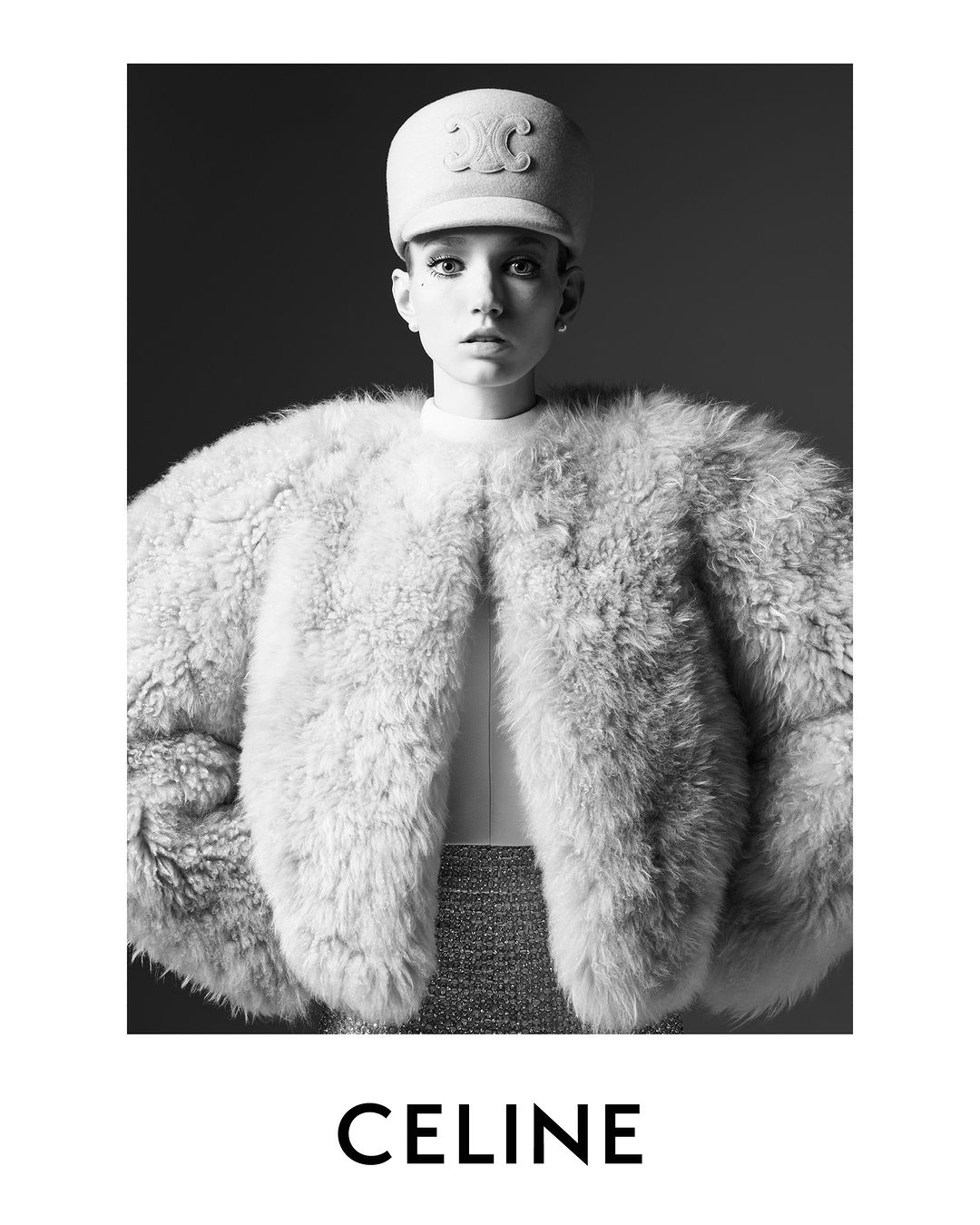 Black-and-white fashion portrait of a young woman wearing a Celine hat and furry jacket, looking at the camera against a dark backdrop.
