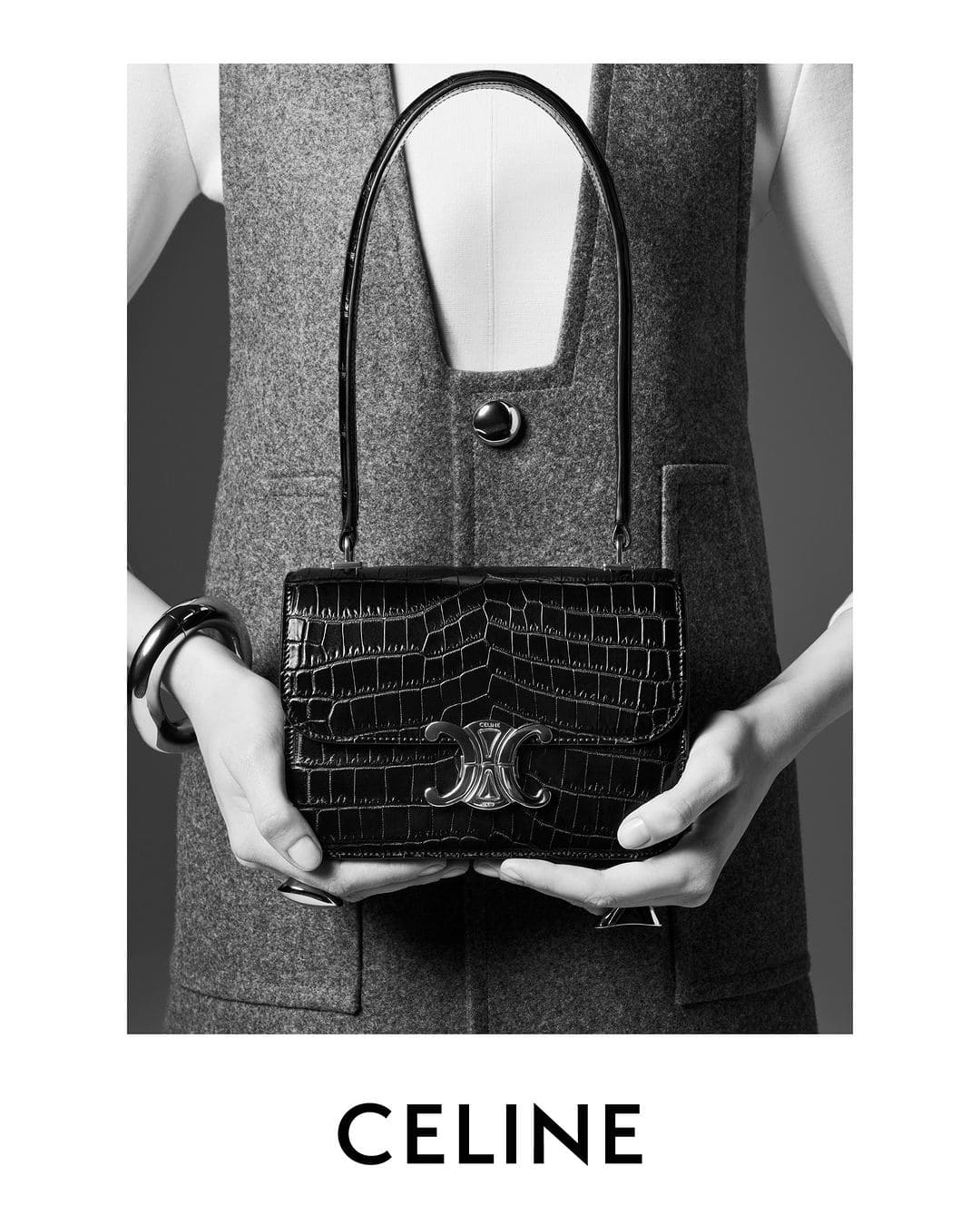 Close-up of a black CELINE purse held in front of a model wearing a grey vest and white blouse. Fashion advertisement in black and white.