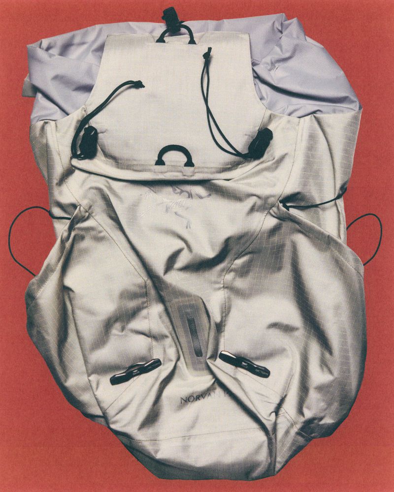 Open grey nylon backpack showcasing its inner compartments and drawstrings, set against a red background in a studio setting.