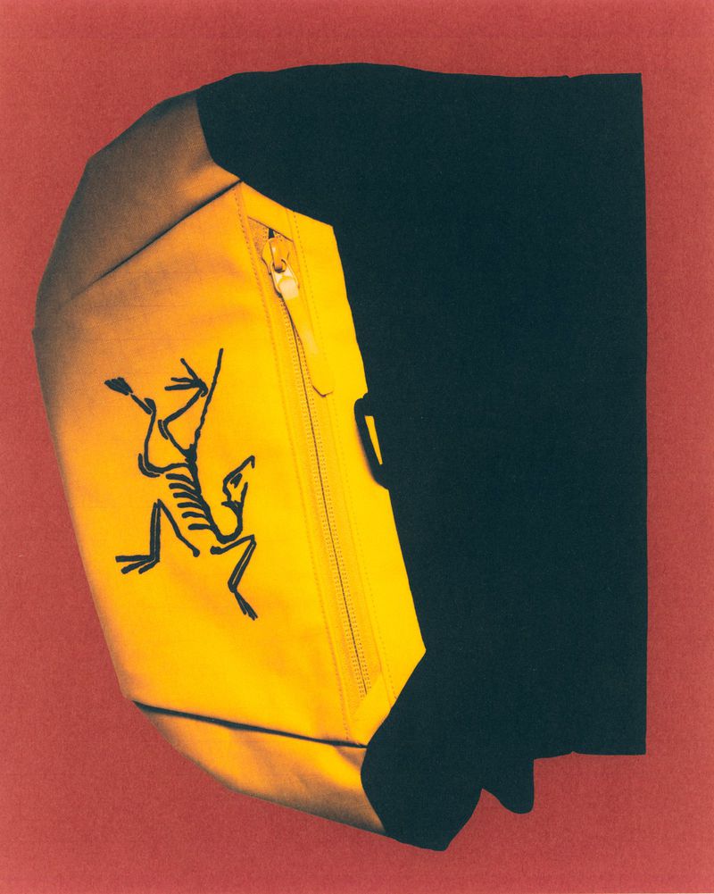 Yellow bag featuring an Arc'teryx logo placed against a background with black fabric on one side and a red wall on the other.
