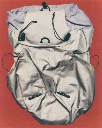 Close-up photograph of a light grey technical backpack with black drawstrings against a textured red background