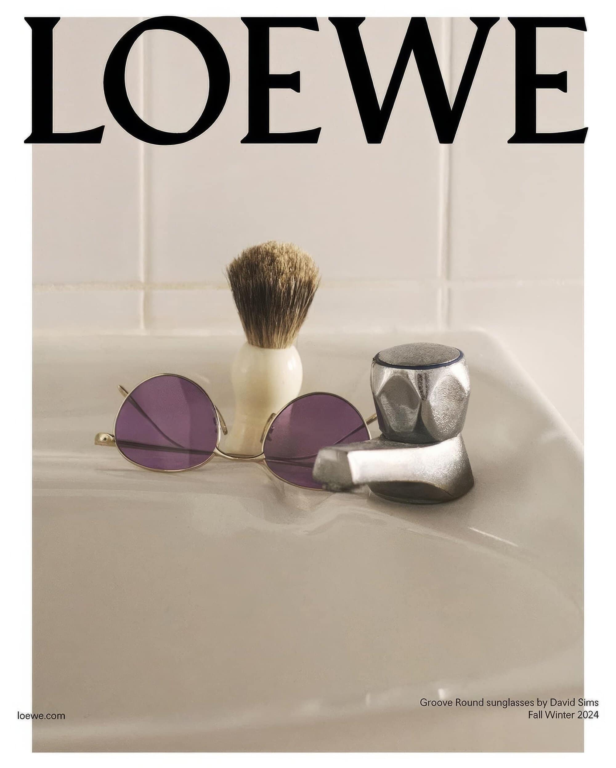 Loewe brand sunglasses with purple lenses showcased alongside a shaving brush and faucet against a cream-colored tiled backdrop.