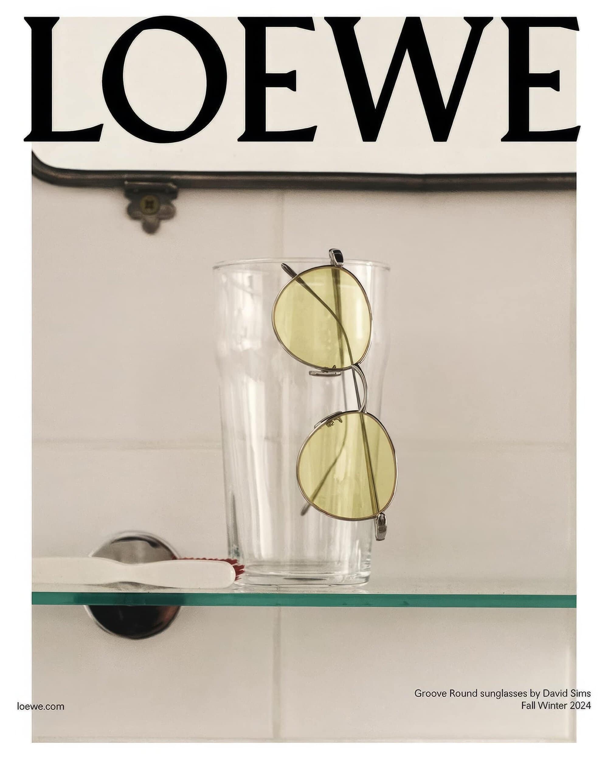 Still life studio shot with yellow round sunglasses on a glass and a toothbrush with Loewe logo signage in the background.