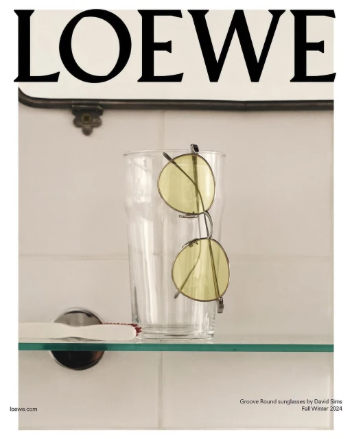 Still life studio shot with yellow round sunglasses on a glass and a toothbrush with Loewe logo signage in the background.