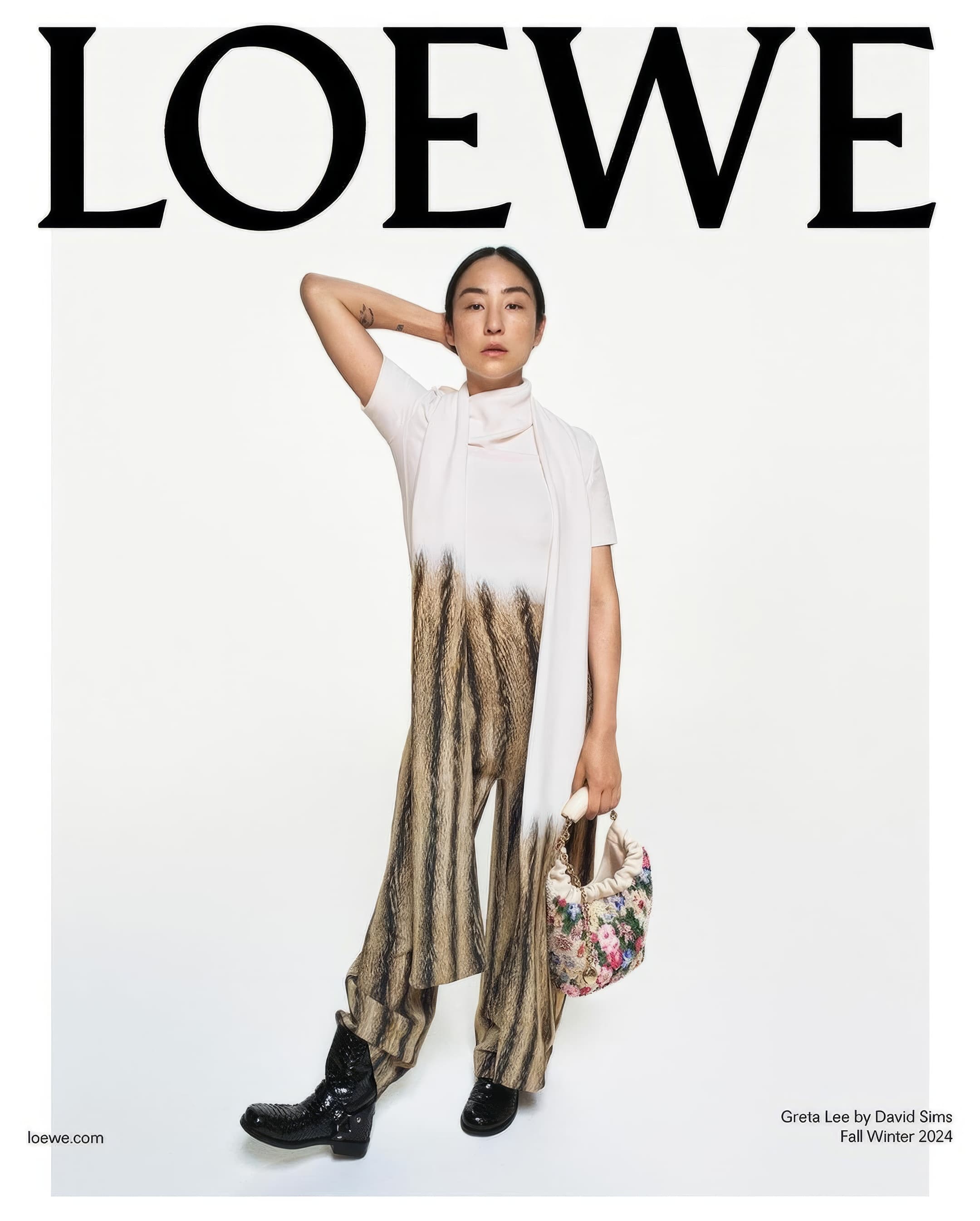 Loewe ad featuring a model in a white top, striped pants, and black boots, holding a floral bag, against a white background.