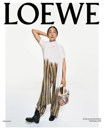 Loewe ad featuring a model in a white top, striped pants, and black boots, holding a floral bag, against a white background.