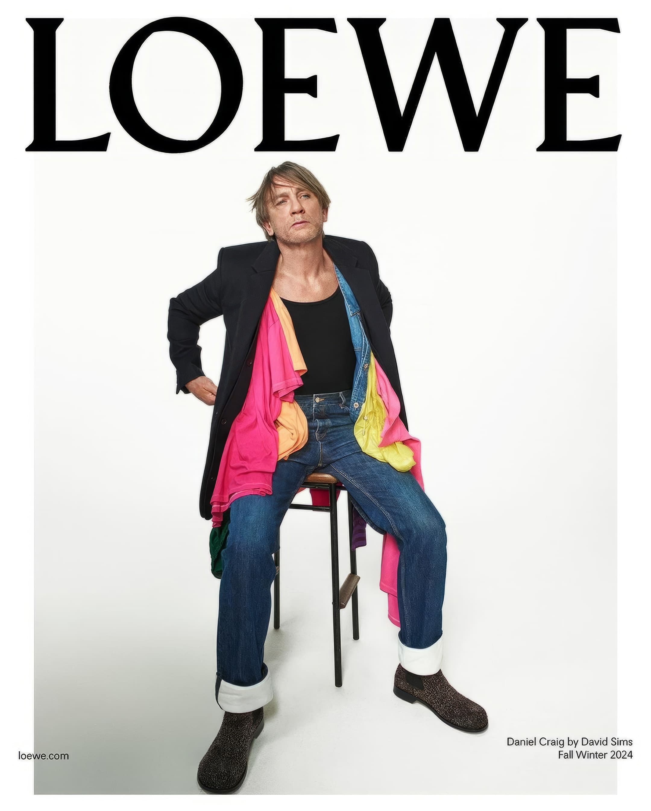 Fashion editorial image of Daniel Craig wearing a black coat over colorful clothes, sitting on a stool against a clean white background.