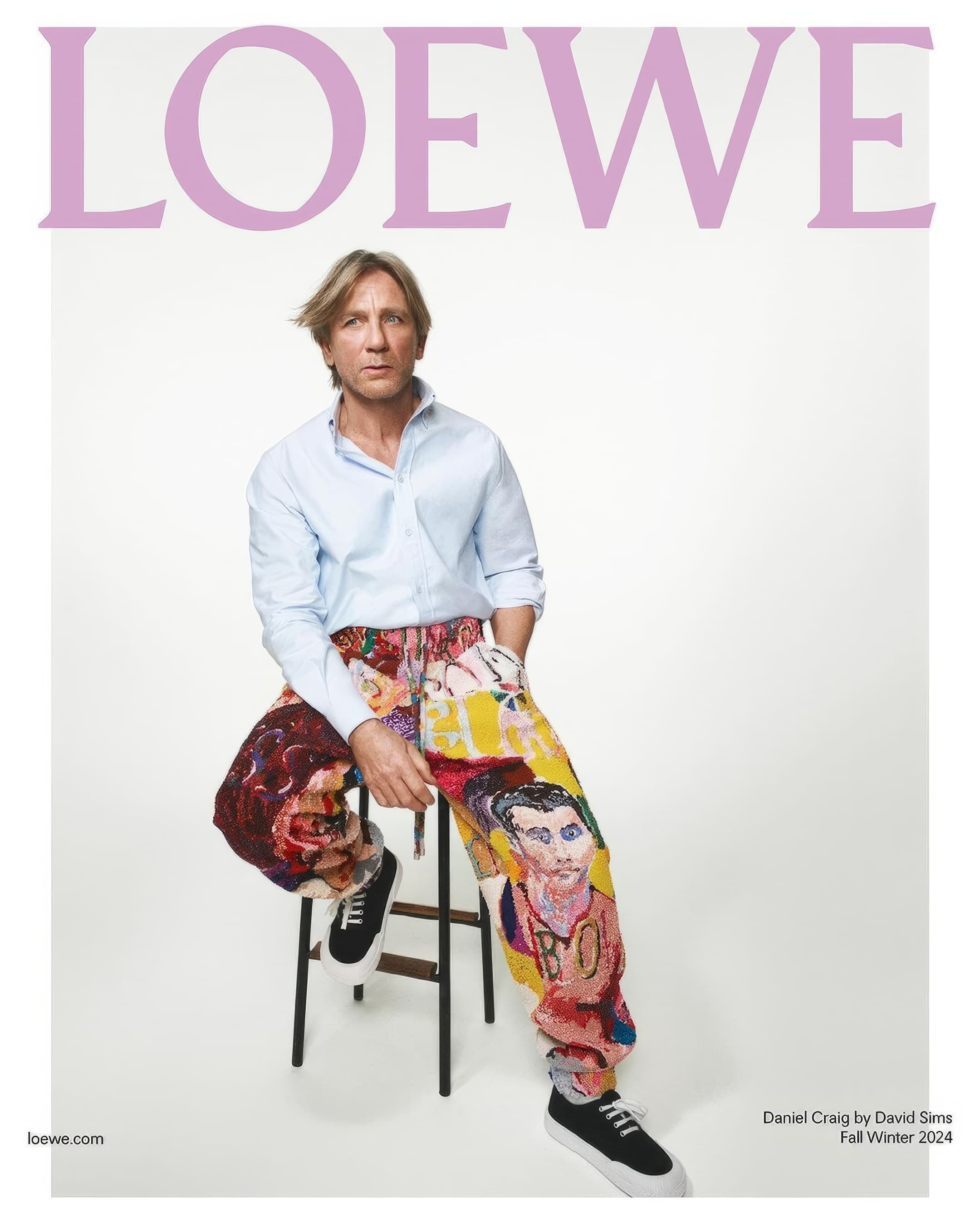 Daniel Craig poses for a Loewe ad, seated on a stool in a studio. He wears an unbuttoned shirt and graphic, colorful pants.