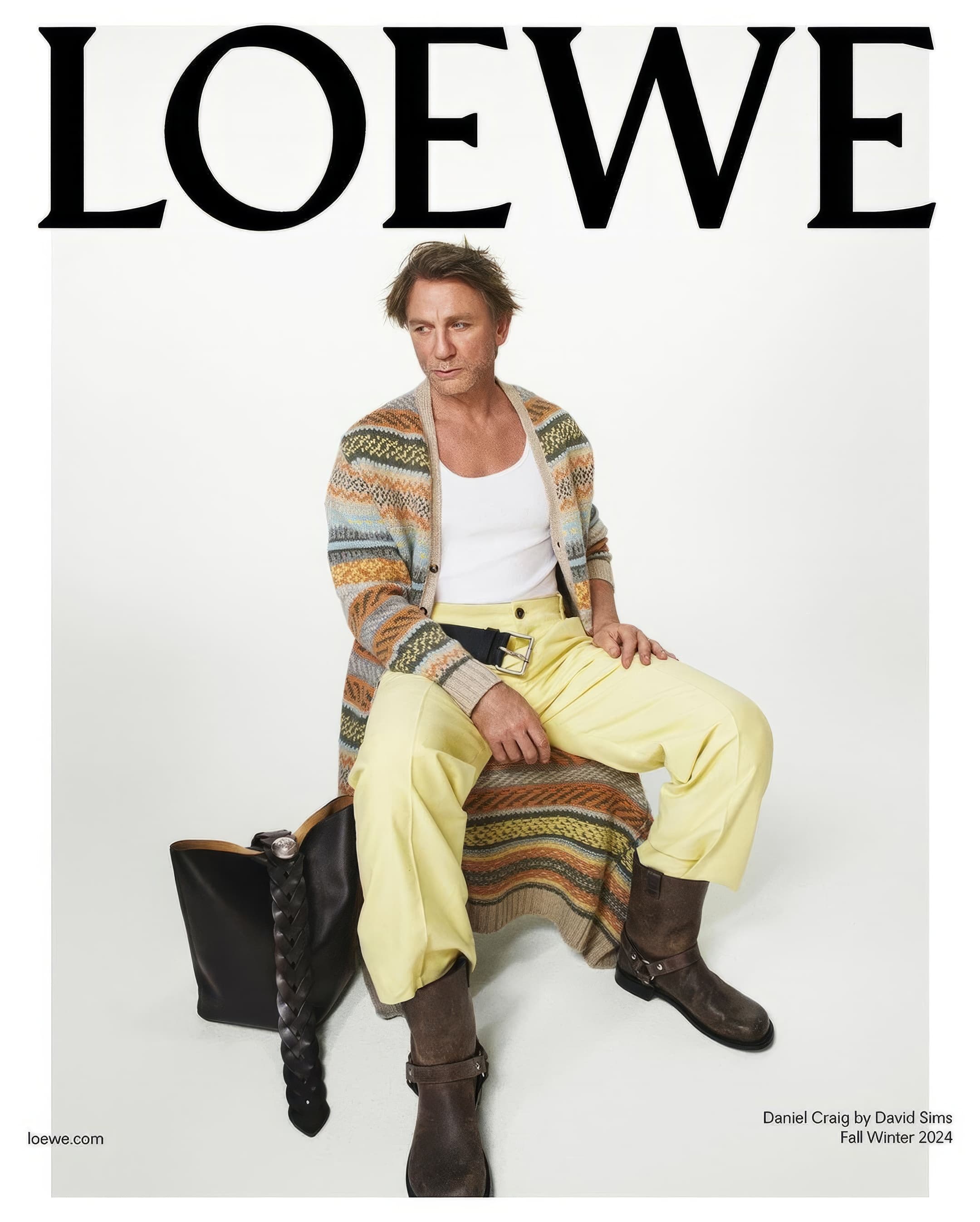 Fashion shot of Daniel Craig sitting on a black leather bag. He is wearing a white tank top, patterned cardigan, yellow pants, and brown boots.