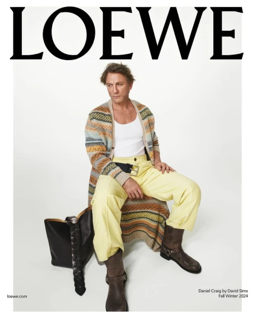 Fashion shot of Daniel Craig sitting on a black leather bag. He is wearing a white tank top, patterned cardigan, yellow pants, and brown boots.