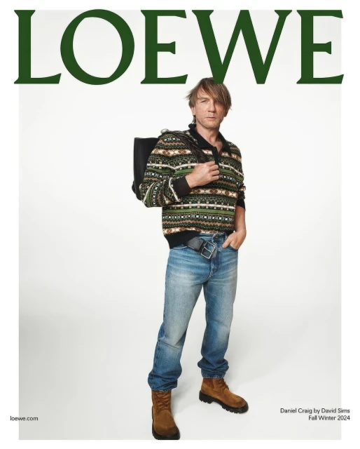 Daniel Craig poses in a Loewe fashion editorial wearing a patterned sweater, jeans, and boots against a clean white backdrop.