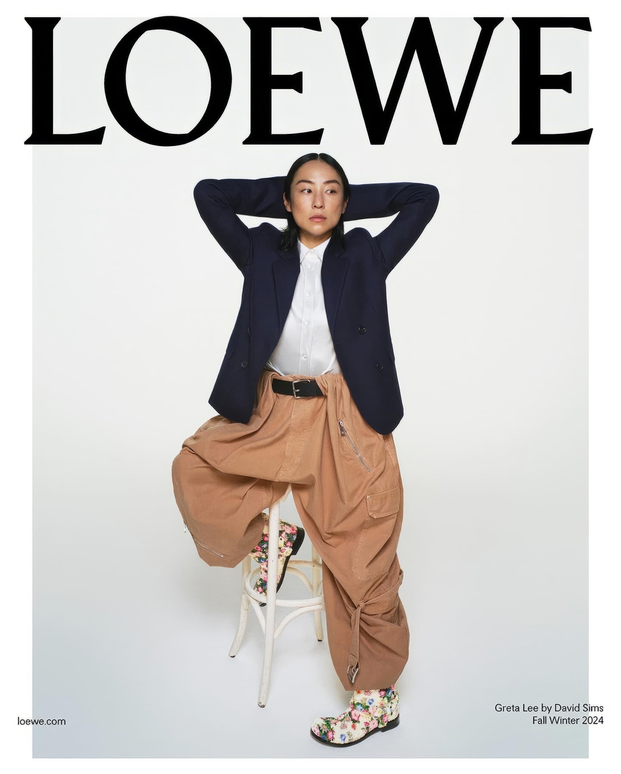 Fashion editorial shot of an Asian woman in a navy blazer and tan, oversized pants, posing on a stool against a white background.