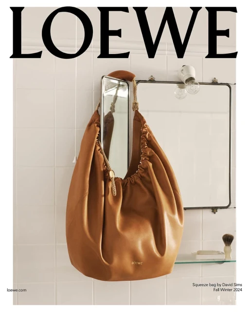 Loewe Squeeze bag in tan leather hangs on a tiled wall next to a mirror and light fixture in a fashion product studio shot.