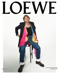 Daniel Craig seated on a stool wearing a dark jacket, layered colorful garments, and blue jeans against a white backdrop.