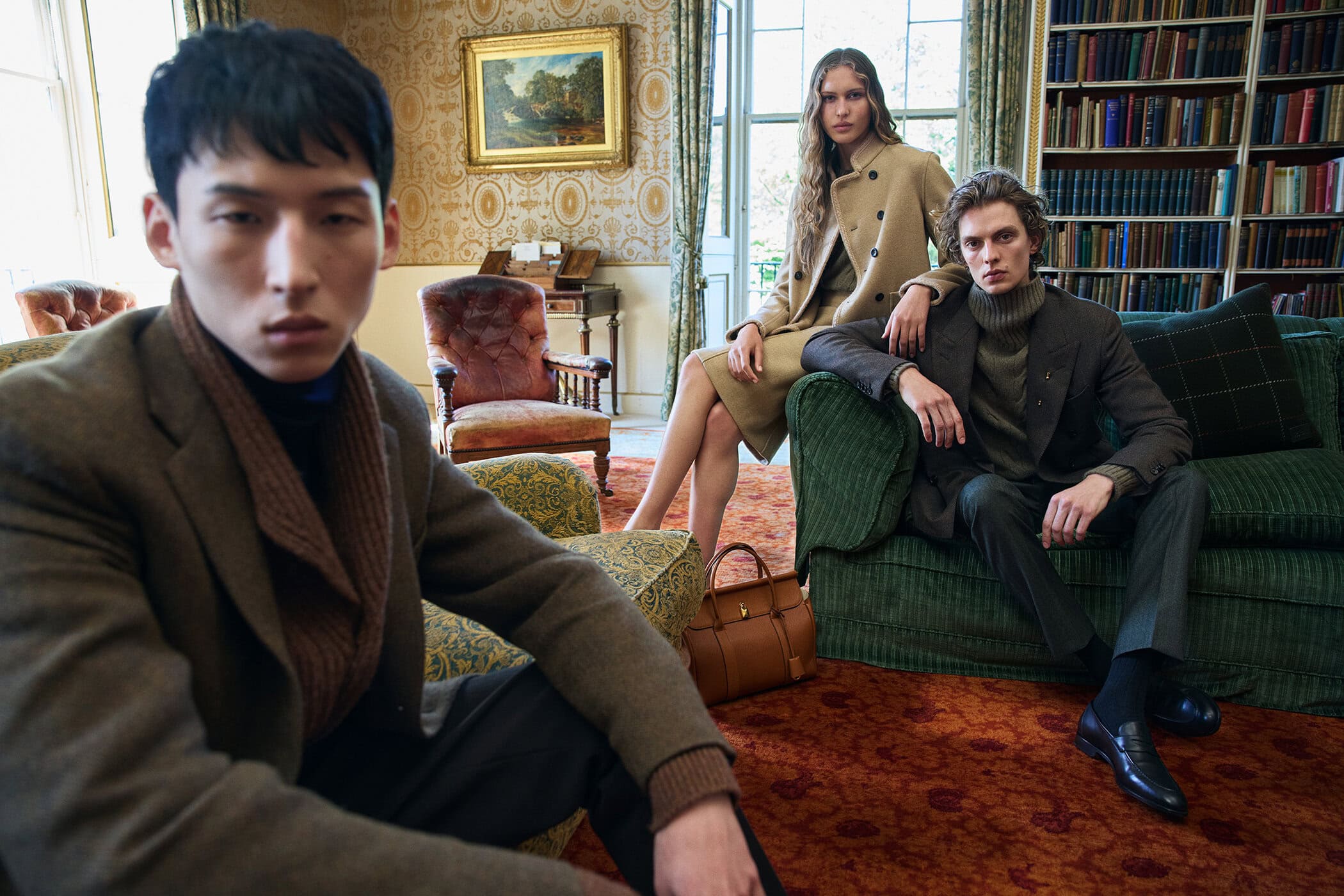 Three fashion models pose in a library setting, showcasing stylish wool coats and blazers in warm tones. Indoor, classic interior.