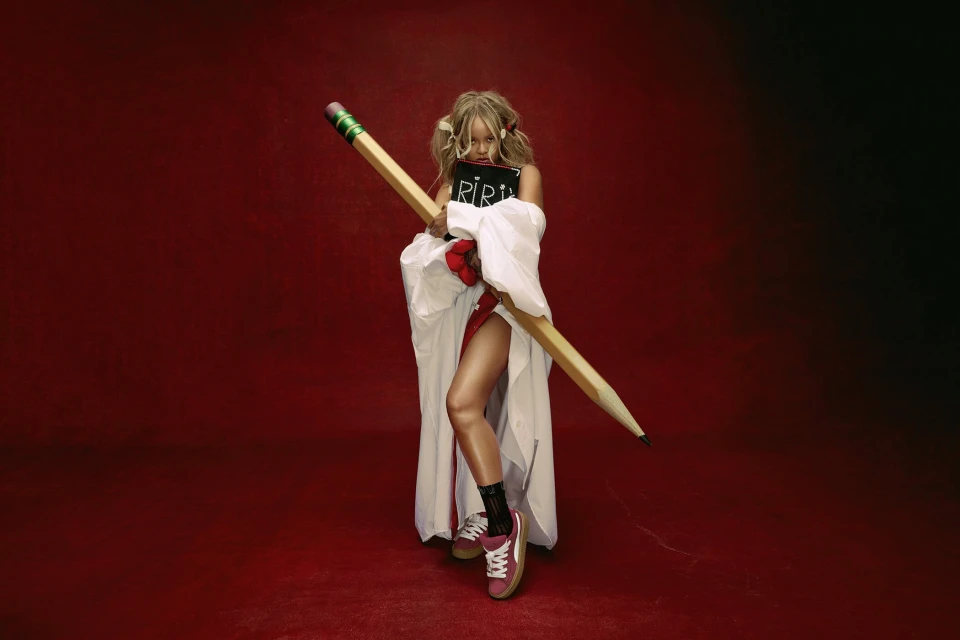 Woman in white coat holds oversized pencil and book in front of a red background in a studio shot with medium framing.