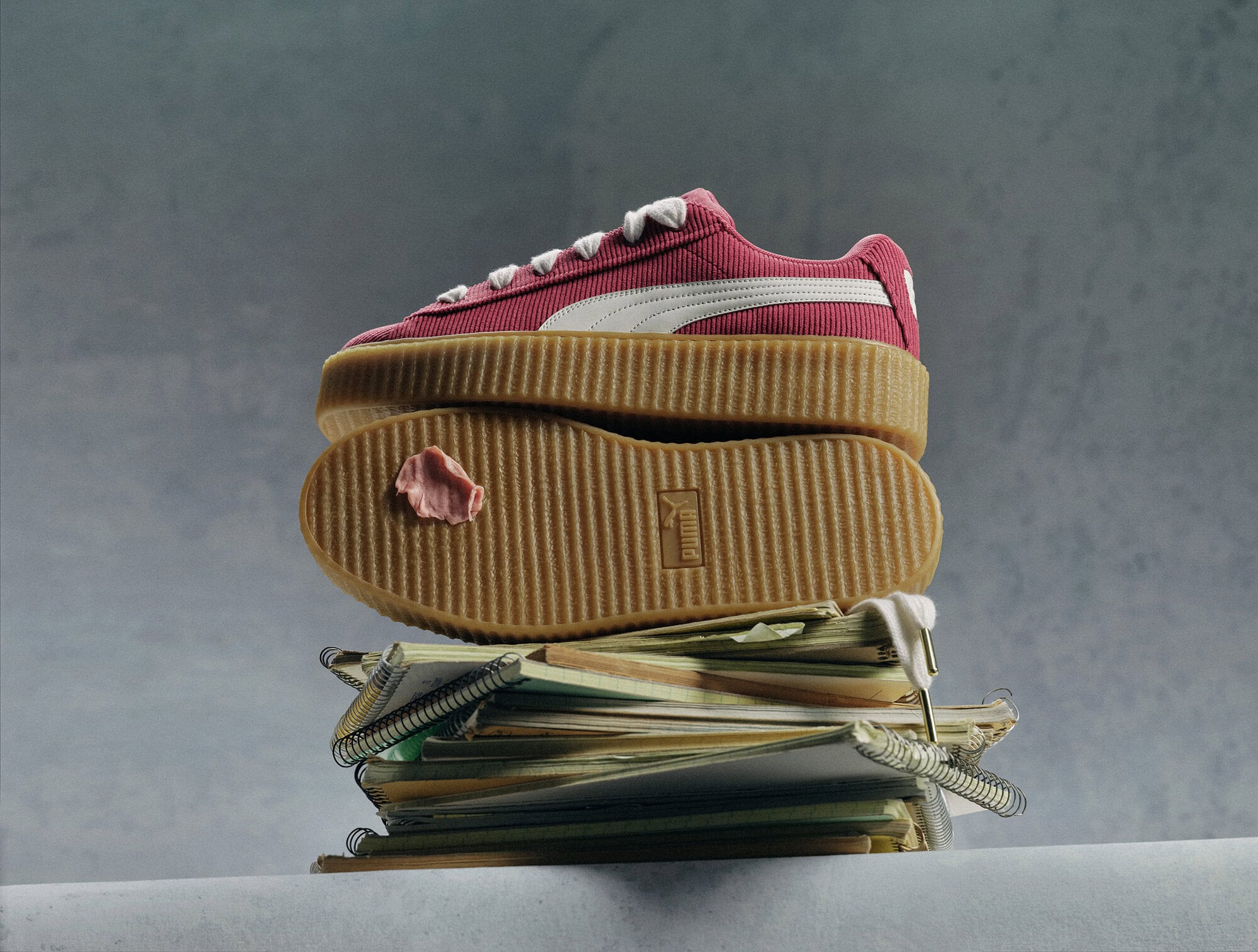 Pink Puma sneaker displayed on top of a stack of spiral-bound notebooks, set against a soft grey background, showcasing a creative studio aesthetic.