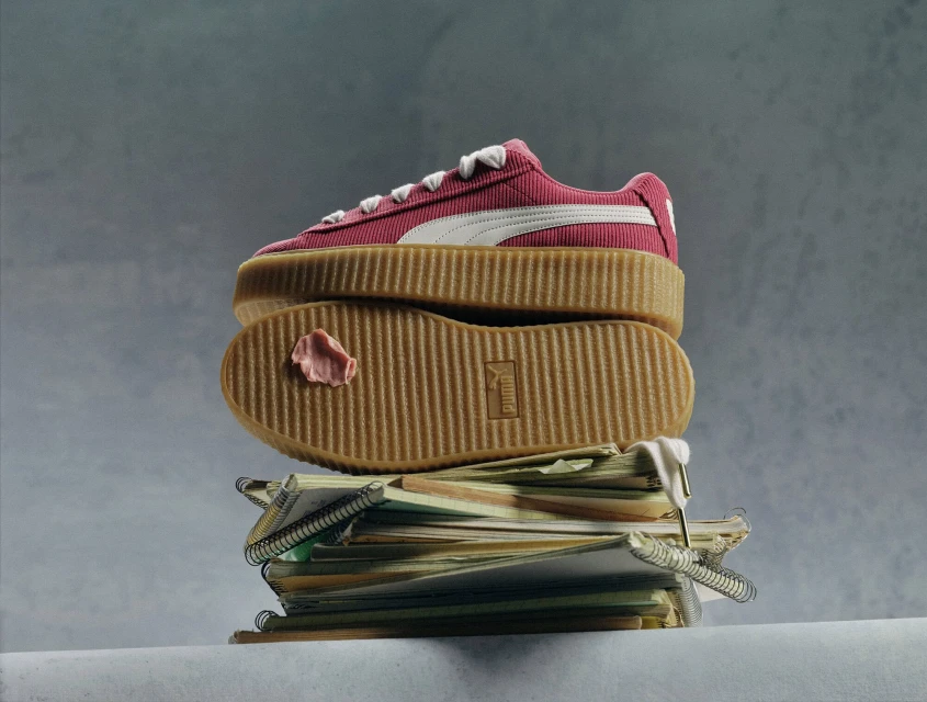 Pink Puma sneaker displayed on top of a stack of spiral-bound notebooks, set against a soft grey background, showcasing a creative studio aesthetic.