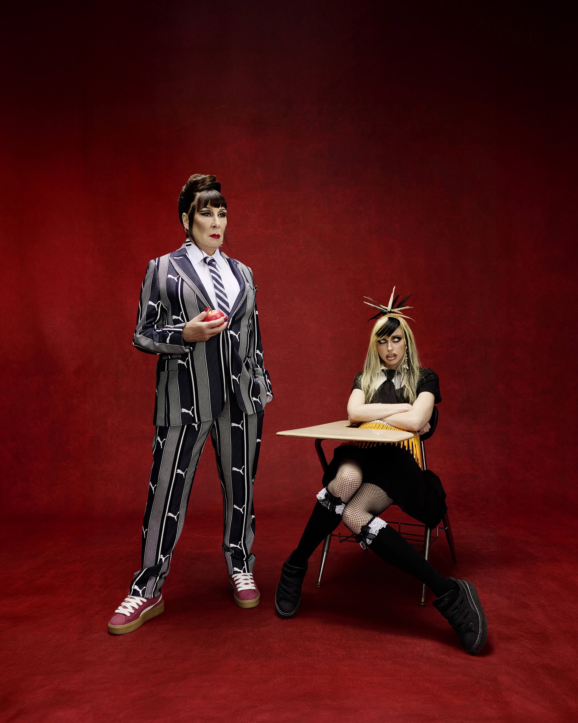 Two people pose against a red background. One in a striped suit holds an apple, and the other in a black dress sits at a desk.
